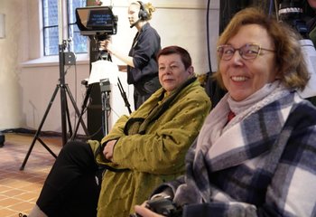 Zwei Frauen sitzend in einem Kirchenraum. Im Hintergrund ein Teleprompter und eine Frau mit Kopfhörern stehend. - Copyright: Annamaria Benckert