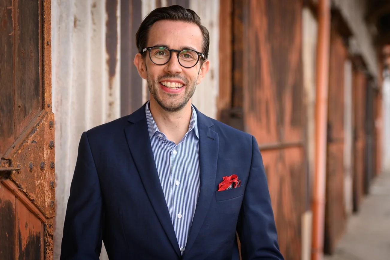 Ein Mann mit Brille, dunklen Haaren schaut freundlich in die Kamera. Er trägt ein Hemd unter einem dunkelblauen Jacket. In der Brusttasche leuchtet ein rotes Einstecktuch. Im Hintergrund Gebäude.