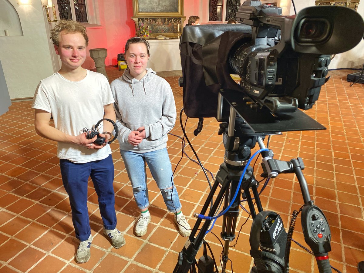 Zwei junge Menschen stehen neben einer Kamera. 