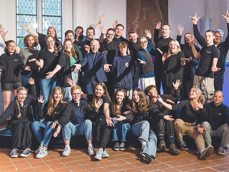 Eine Gruppe von Menschen stehen und sitzen hintereinander, einige haben die Arme hochgestreckt